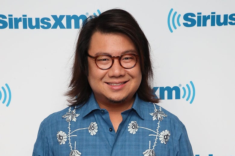 Kevin Kwan, wearing a blue shirt with white flowers and black glasses, stands and smiles at the camera.